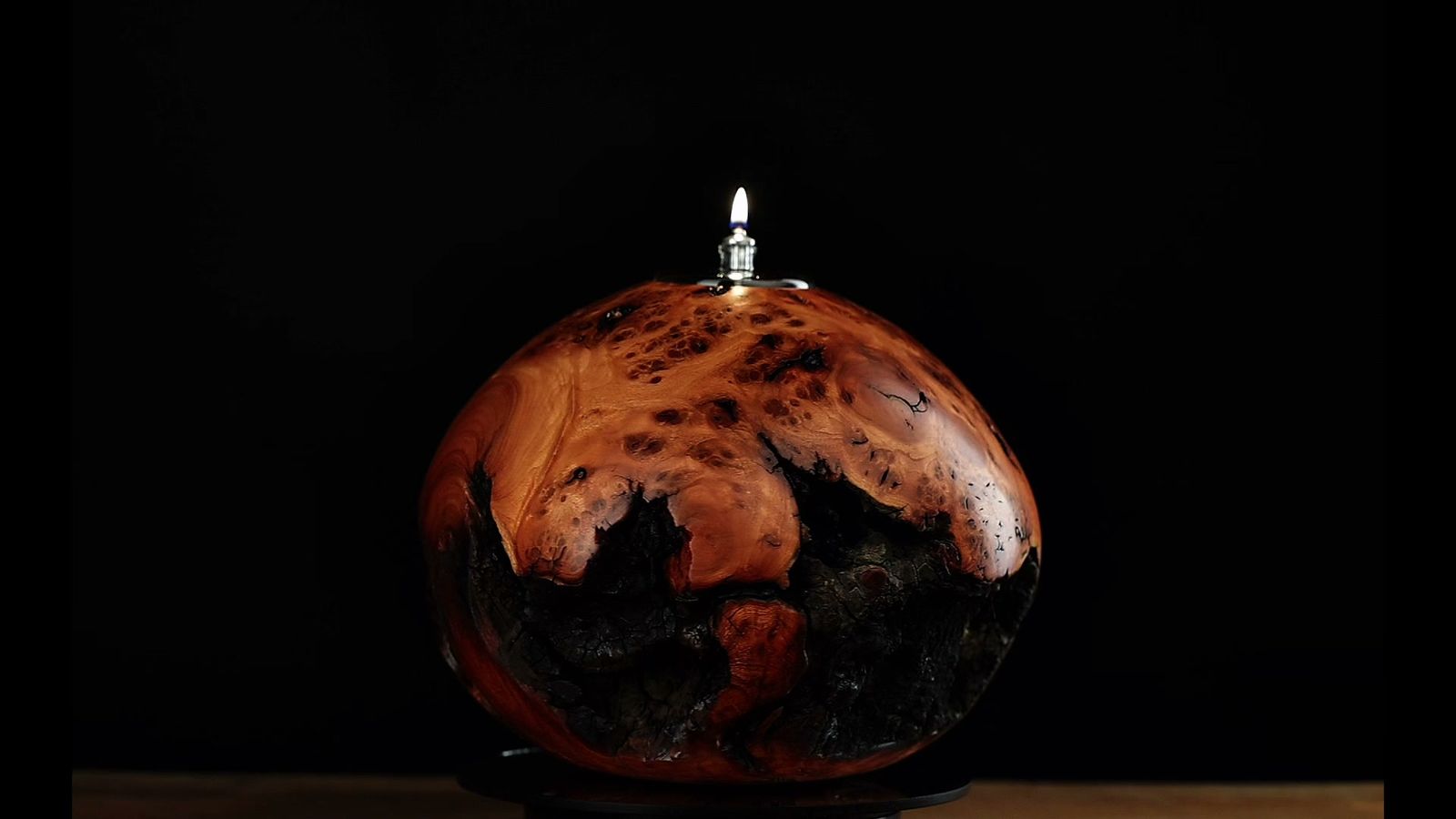 A hand-turned juniper burl oil lamp, lit, on a wooden table against black
